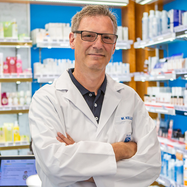 Apotheker Keller in der Apotheke Apotheker Keller steht mit verschränkten Armen vor Regalen voller Medikamente und Kosmetika im hellen Apothekeninnenraum.
