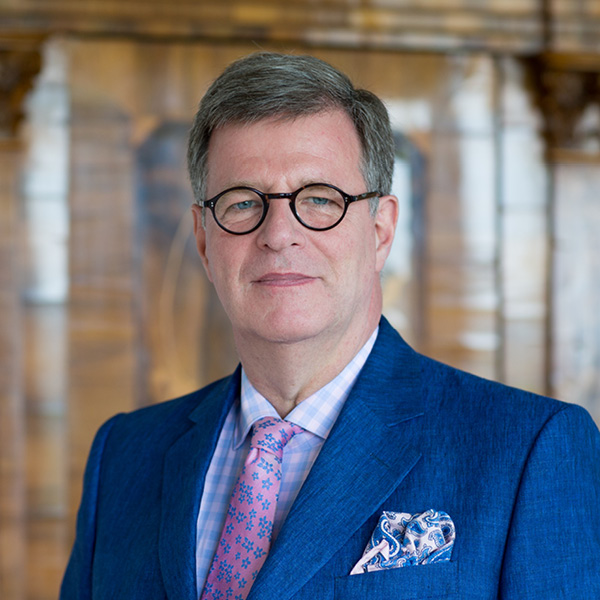 Joerg Geller in seinem Büro Joerg Geller in einem blauen Anzug mit rosa Hemd und Krawatte vor einer historischen Kulisse stehend.