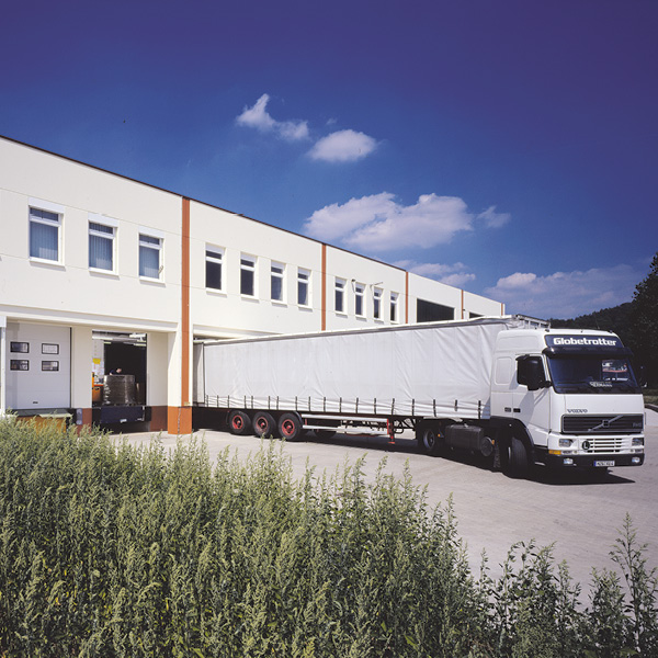 Logistikzentrum mit LKW Logistikzentrum kohlpharma Merzig mit LKW vor einer Lagerhalle unter blauem Himmel und grunen Straeuchern im Vordergrund.