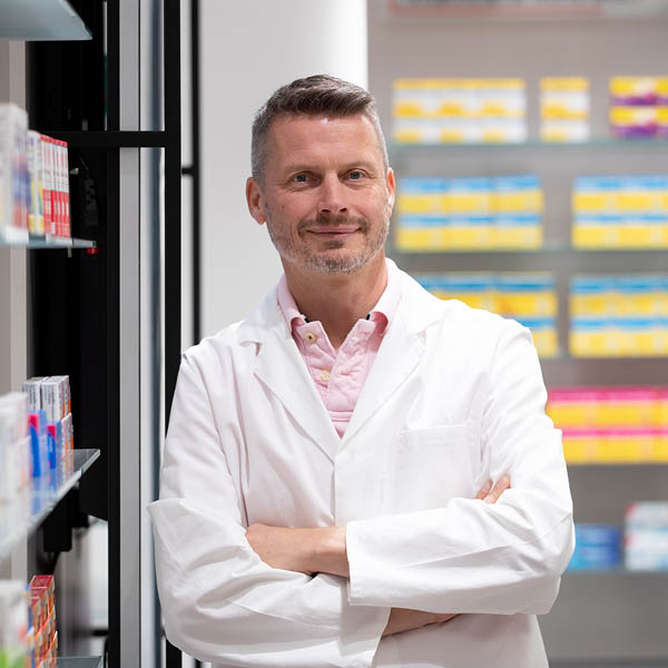 Apotheker in der Offizin Apotheker steht mit verschränkten Armen vor Regalen voller Medikamente und lächelt freundlich in die Kamera.