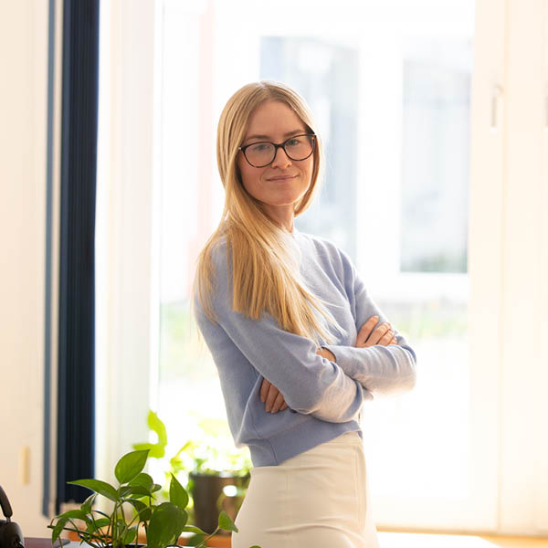 Verwaltung 3 im Büro Verwaltung 3 steht im Raum mit verschränkten Armen und blickt in die Kamera. Sie trägt eine helle Bluse und weiße Hose.
